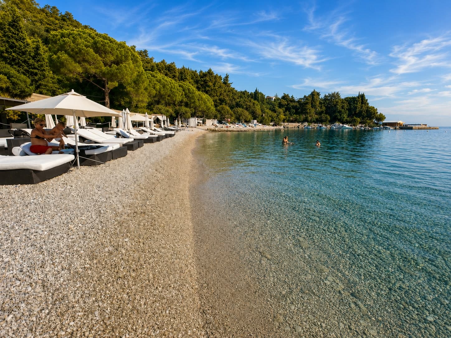 White pebbles and clear shallow water on a beach near Lovran, loungers under the pines